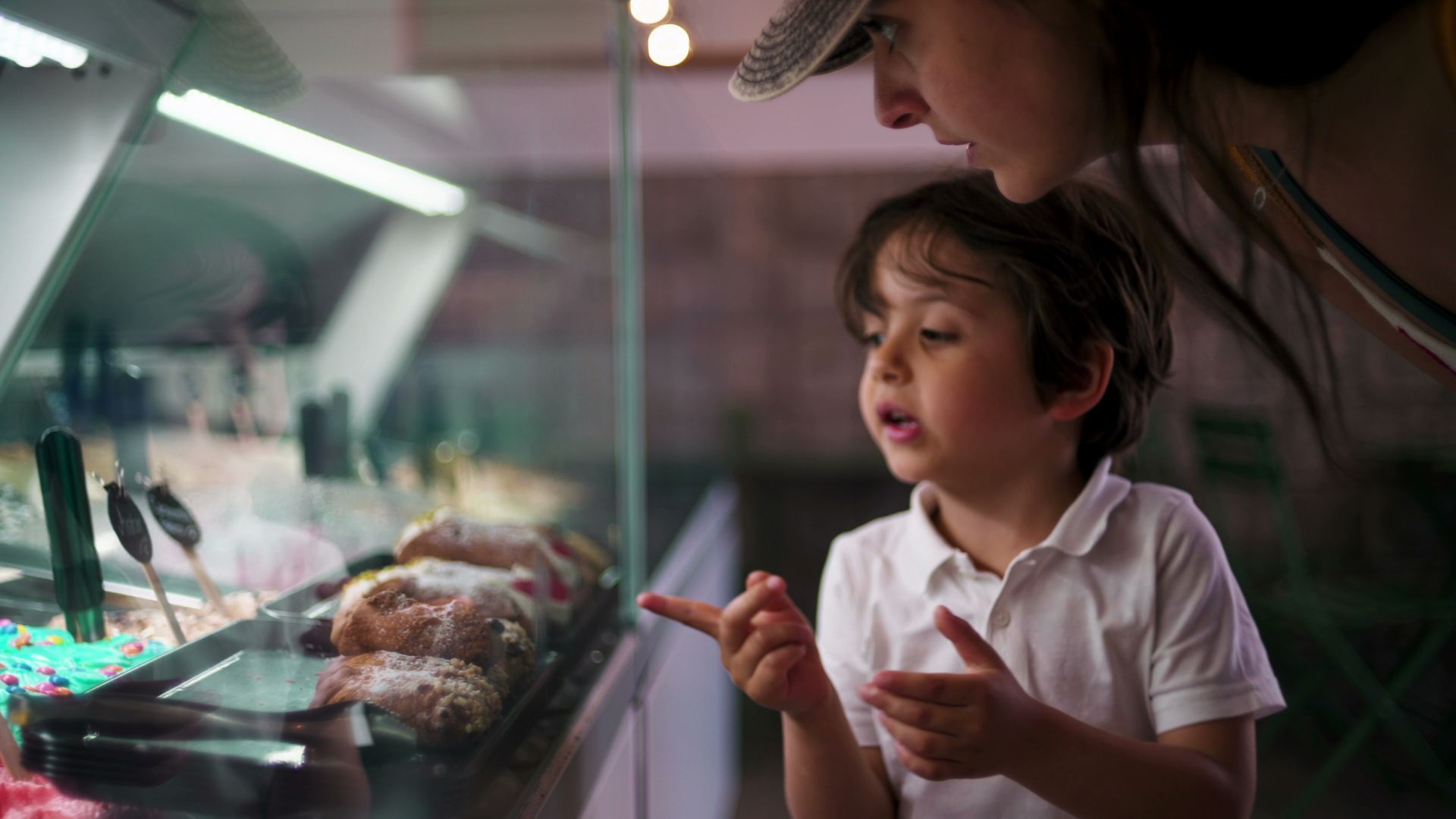 Un garçon désigne du doigt des pâtisseries dans une vitrine, sous le regard d'une personne coiffée d'un chapeau, probablement un gardien.