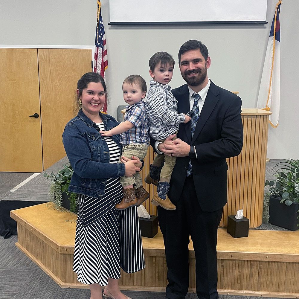 Family of four stands on a wooden stage in a church. Man holds one child, woman holds another.