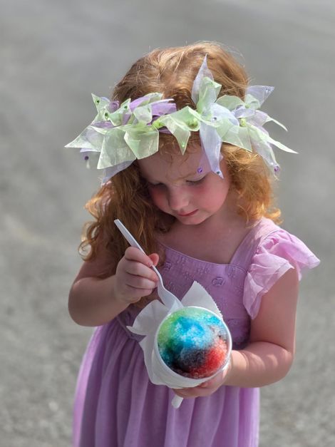 A young girl with red hair and a flower crown enjoys a colorful snow cone