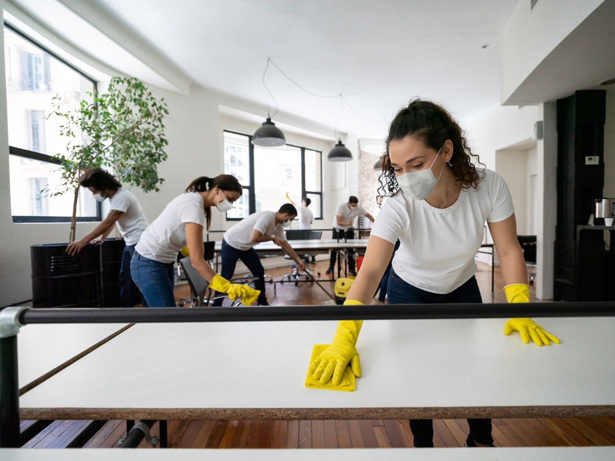 A woman wearing a mask and yellow gloves is cleaning a table in an office.