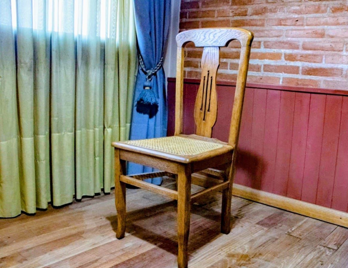 Silla de madera con asiento tejido contra pared de ladrillo, cerca de cortinas.