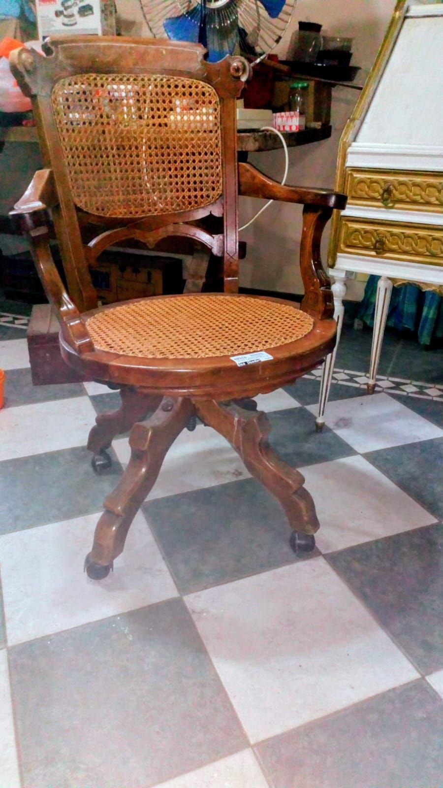 Silla de oficina de madera con asiento y respaldo de caña, diseño ornamentado, sobre suelo de baldosas.