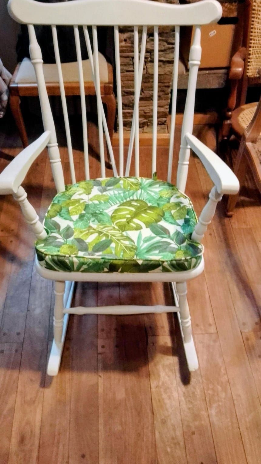 Mecedora de madera blanca con cojín de hojas tropicales verdes.