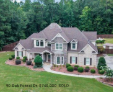 An aerial view of a two-story beige house with a stone facade, situated on a grassy lot surrounded by a dense forest. $740,000, SOLD.