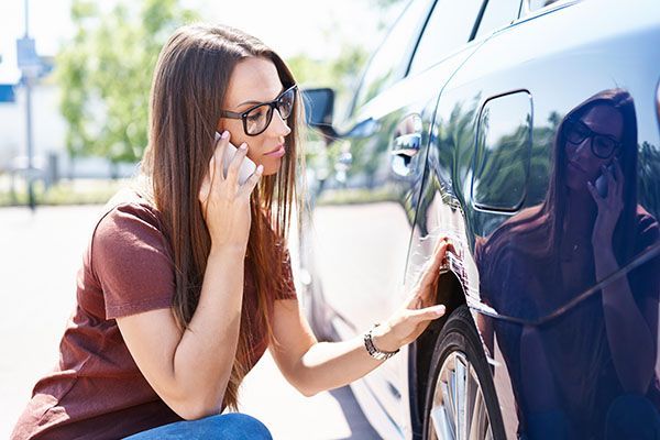 Woman on phone examines damage on car door.