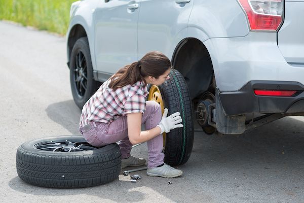 Woman changing a car tire on a roadside, wearing gloves and holding a spare tire. | Auto Clinic Care