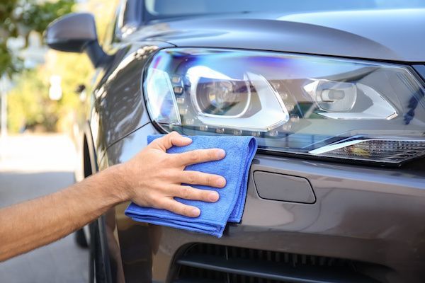 Person wiping a gray car's headlight with a blue microfiber cloth outdoors on a sunny day. | Auto Clinic Care