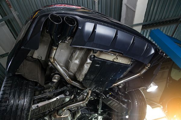 Underside of a car in a garage, exhaust pipes, black bumper, metal components, and tires.