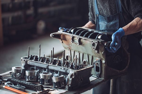 Mechanic holding an engine block, wearing blue gloves and overalls, in a workshop. | Auto Clinic Care