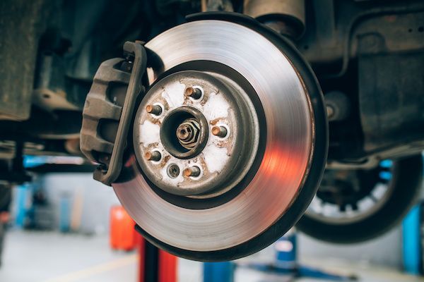 Car's brake disc and caliper in a repair shop. The disc is silver, and the caliper is dark with the car raised.