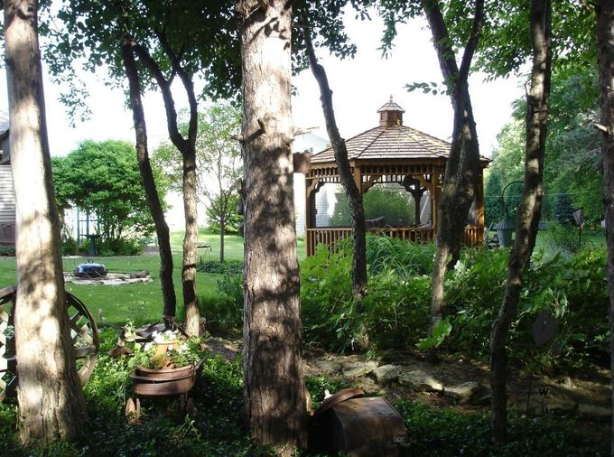 Wooden gazebo in a garden, framed by trees. Green grass and foliage.