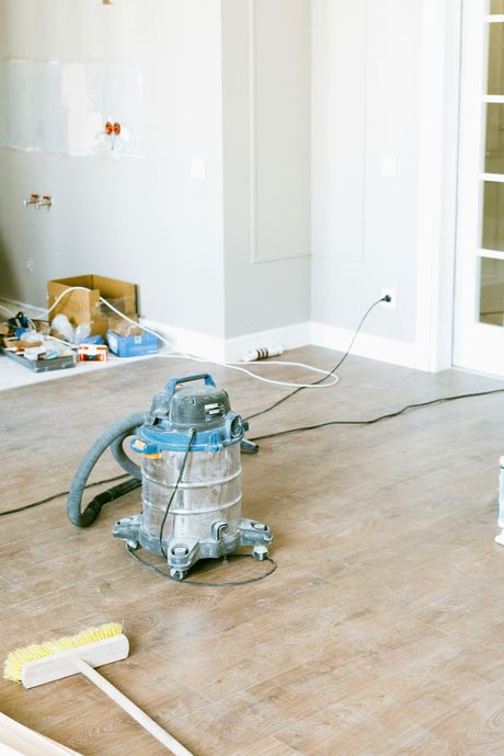 Wet/dry vacuum in a room under construction with wood-look flooring; a broom lies nearby.