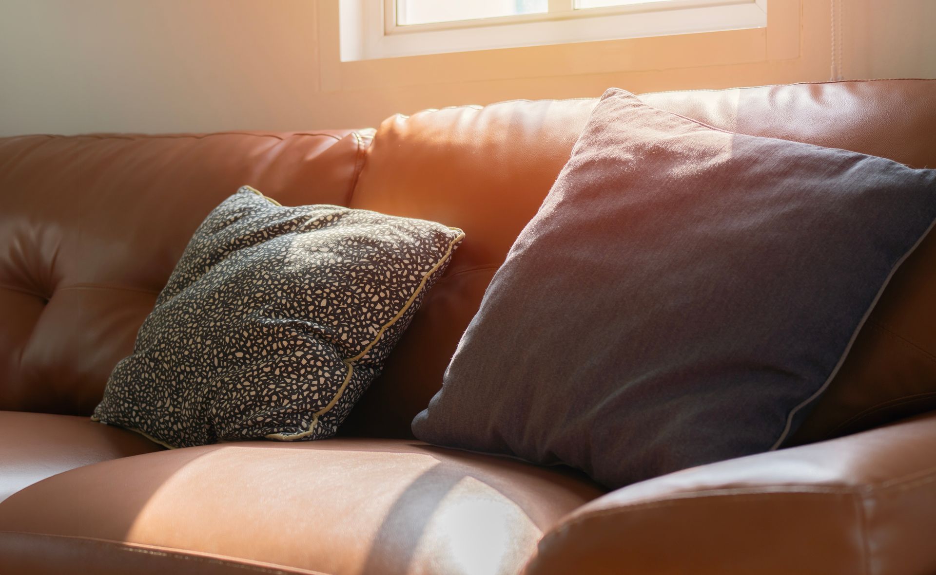 Brown leather couch with two pillows: one dark patterned, one solid blue. Sunlight streams in.