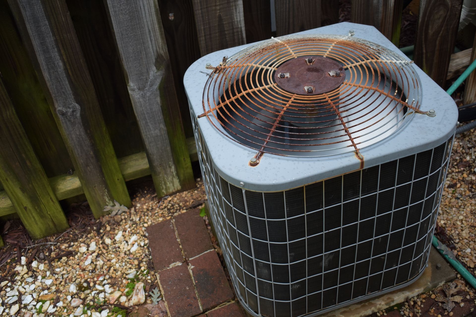 Outdoor air conditioner unit with a rusted fan grill beside a wooden fence and brick pavers