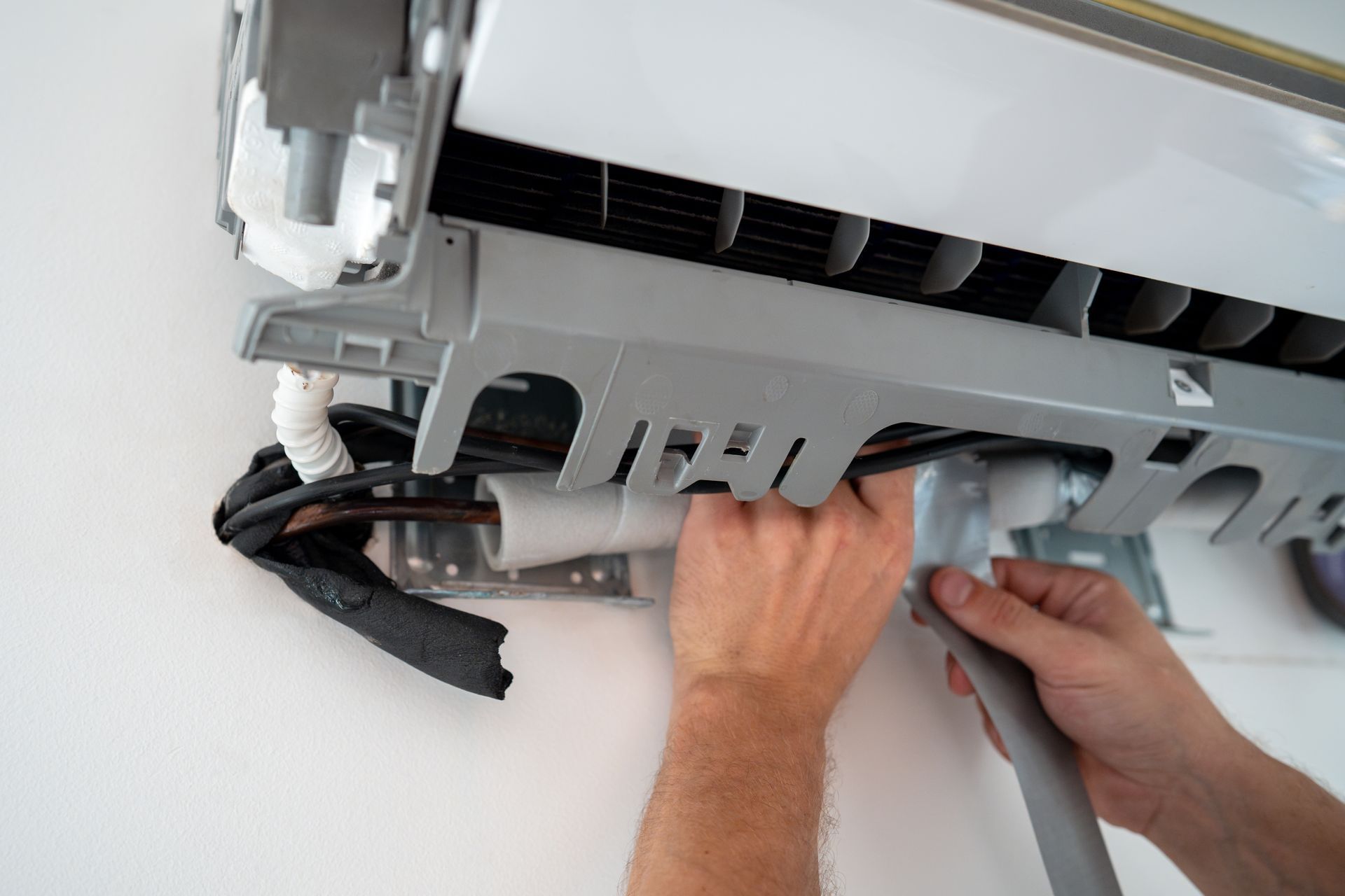Hands installing a wall-mounted air conditioner, with exposed wiring and tools visible.