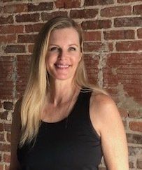 A woman in a black tank top is smiling in front of a brick wall.