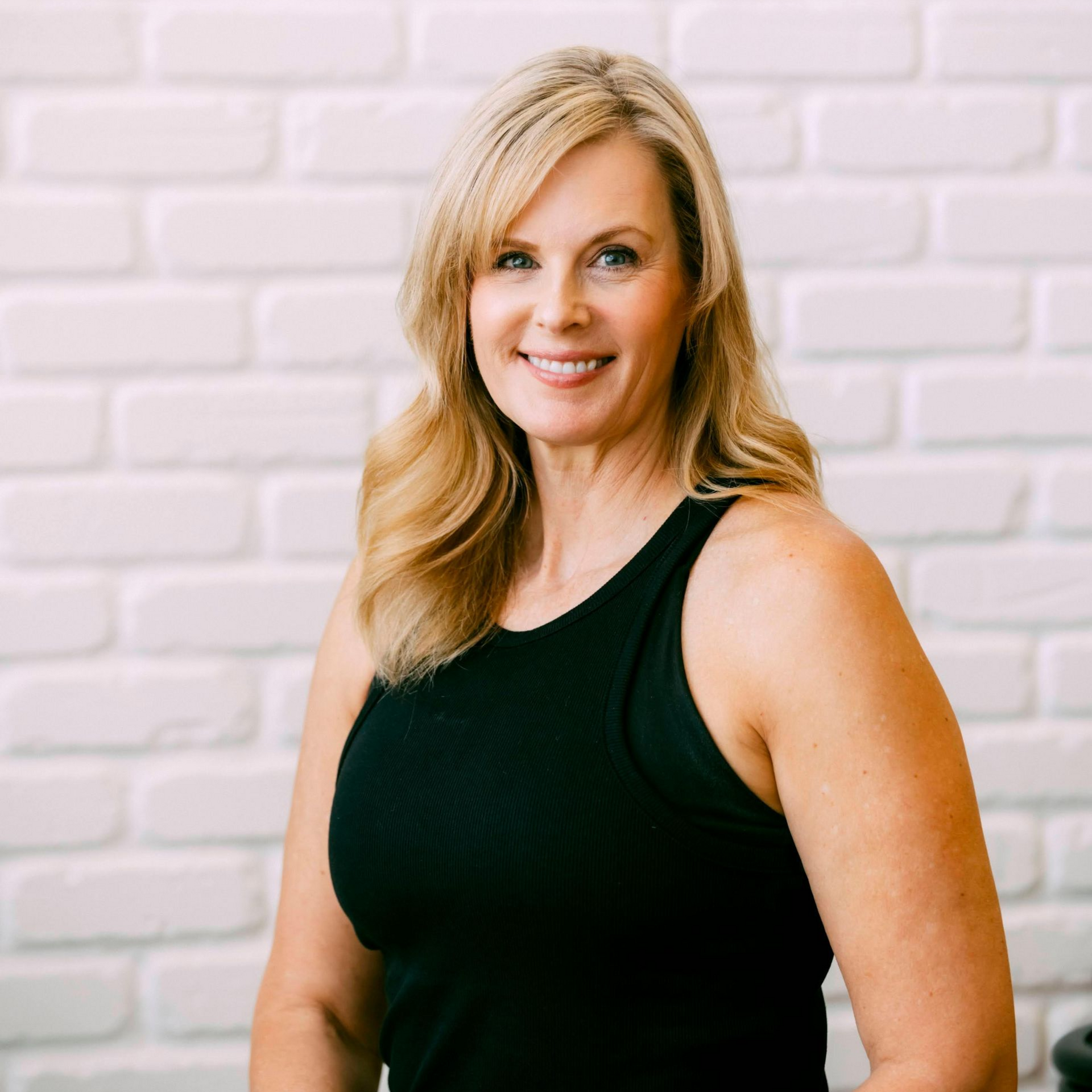 Colette in a black tank top is smiling in front of a brick wall .