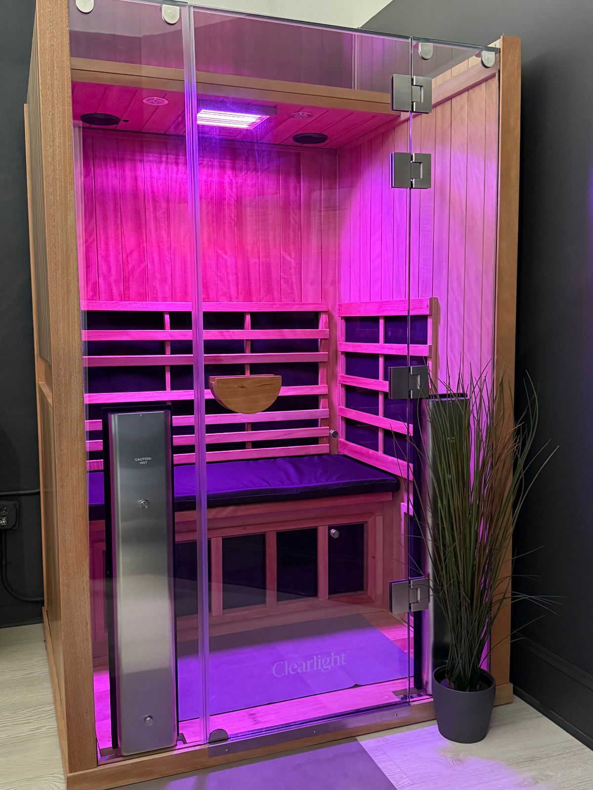 A small, wooden sauna with a glass door, lit with pink light, and a bench inside.