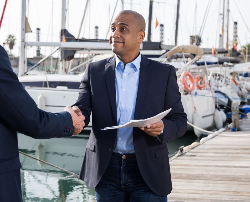 Two men are shaking hands in front of boats in a marina.