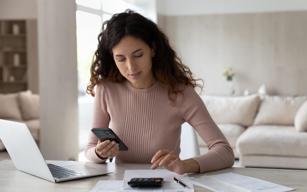 A woman is sitting at a table with a laptop and a cell phone.