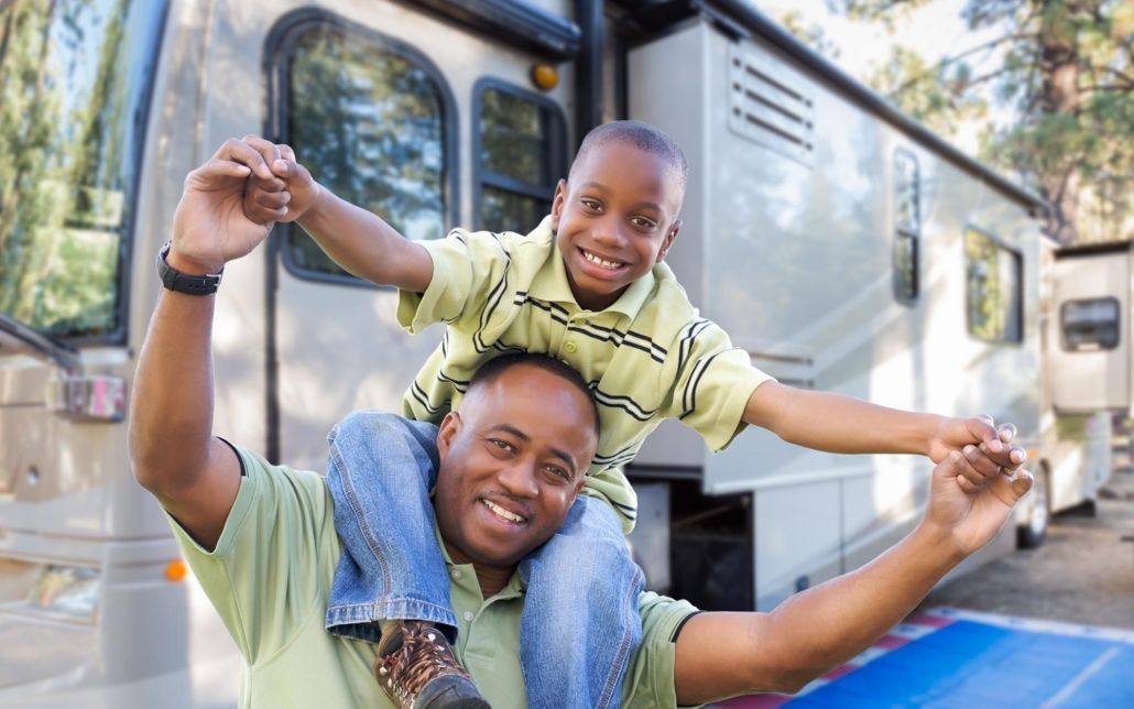 A man is carrying a child on his shoulders in front of a rv.