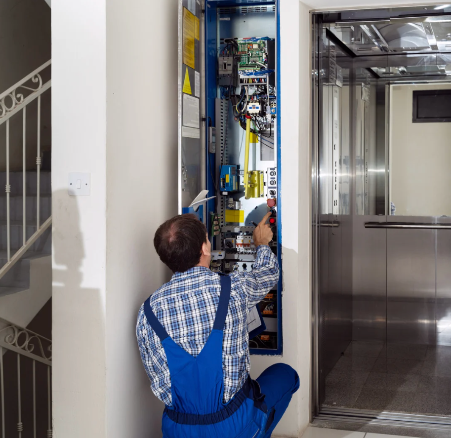 A repairman in blue overalls working on an elevator control panel in a building hallway.