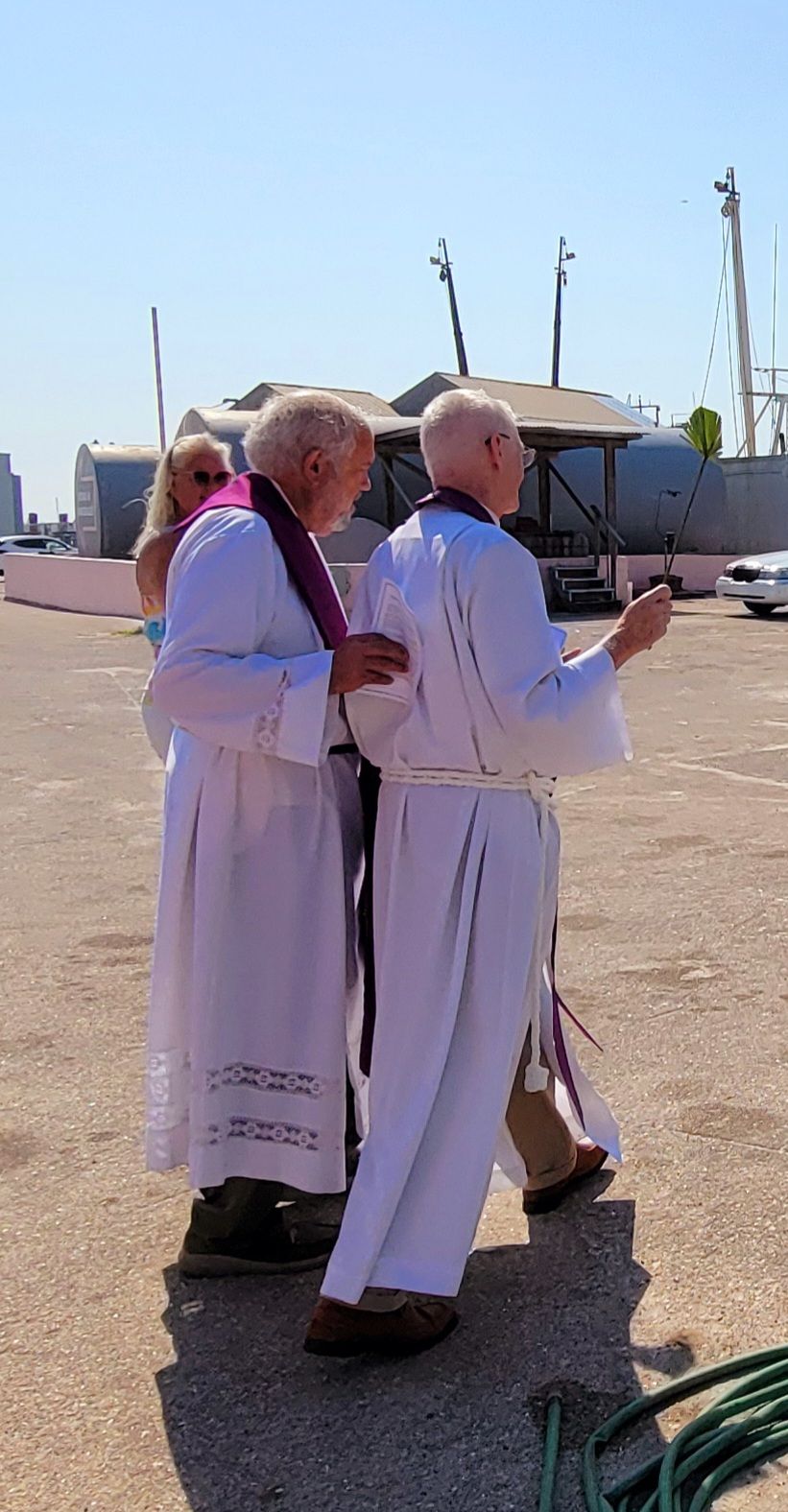 Two priests in white robes with purple stoles, outdoors, appear to be leading a ceremony.