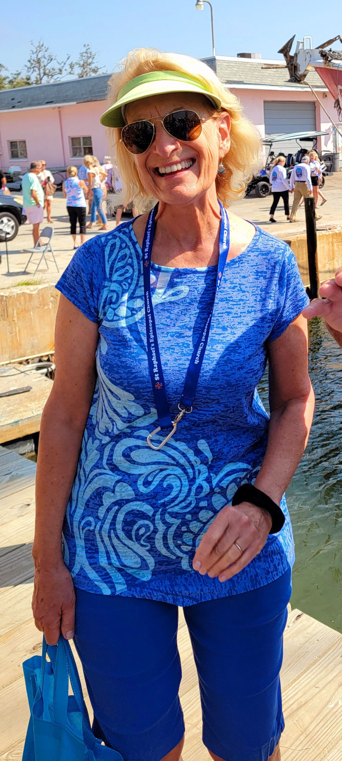 Woman smiles, wears sunglasses, visor, blue shirt, pants, holds bag on pier.