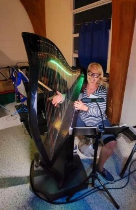 Woman playing a large black harp indoors; smiling. Wooden beams, a microphone, and music stand are visible.