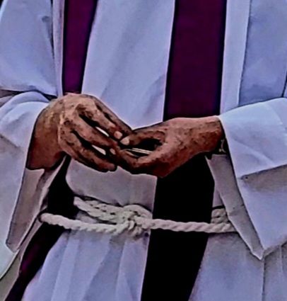 Person in white robe, hands clasped, white rope belt. Purple detail visible on robe.