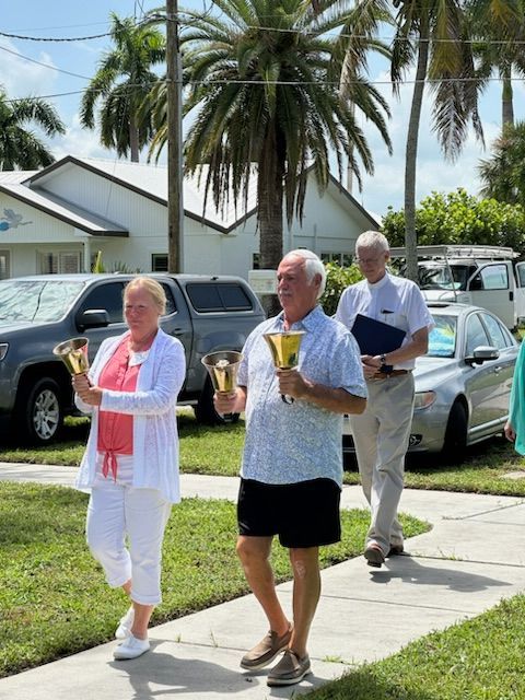 Three people walking on a sidewalk, ringing bells. Light skin, outside, sunny.