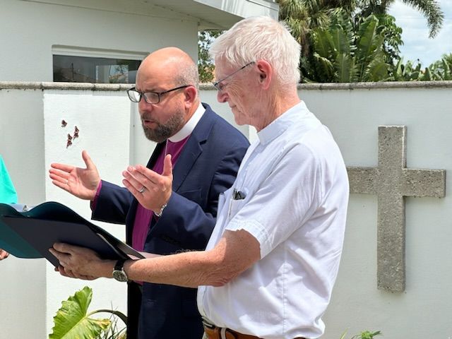 Two men in clerical attire beside a cross. One gestures with hands, reading from a book. White wall background.