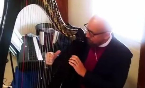 Man in clerical collar playing a harp, smiling, indoors near a window.