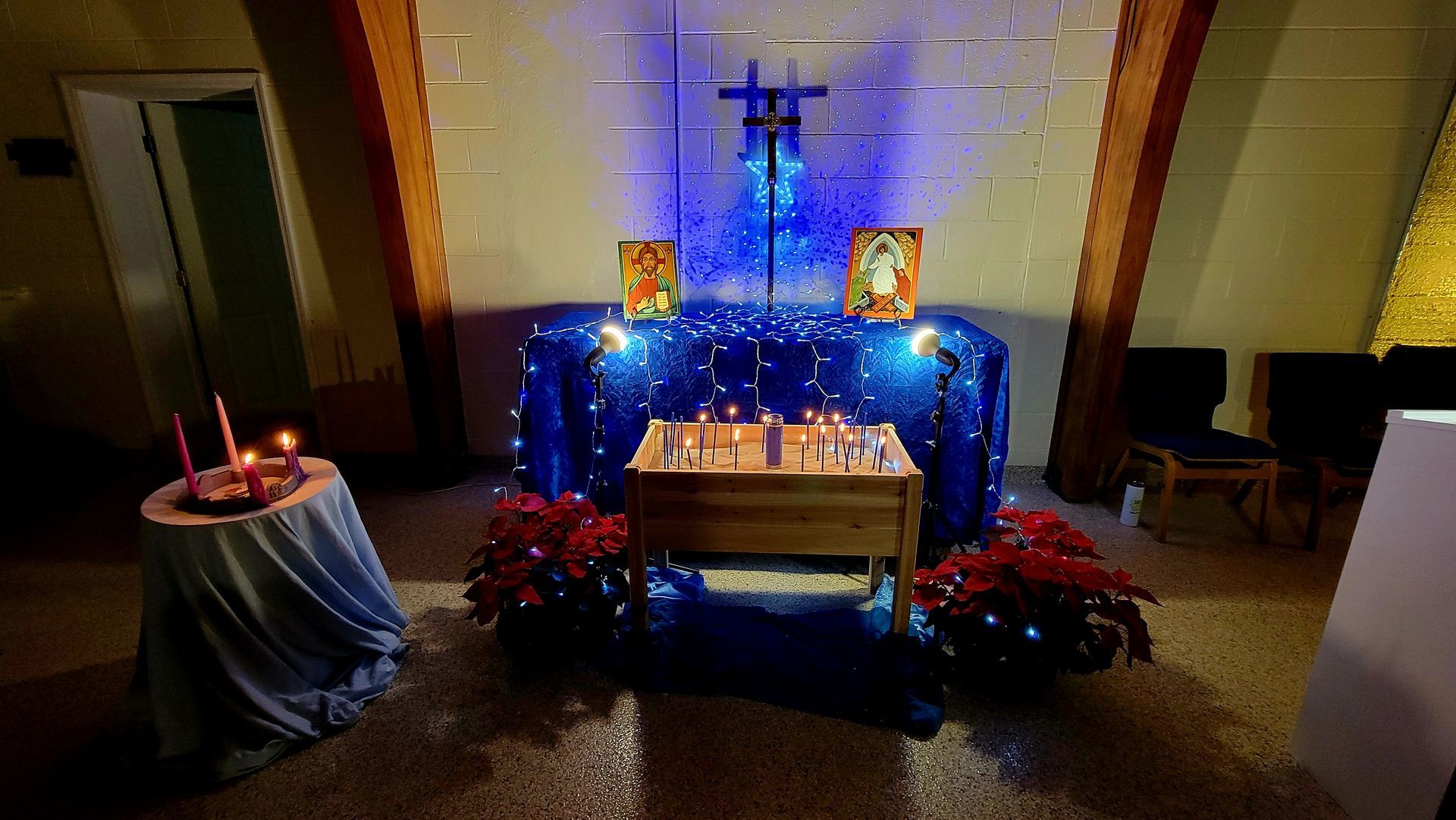 Altar with candles, two icons, and a cross, illuminated with blue light, set against a wall.