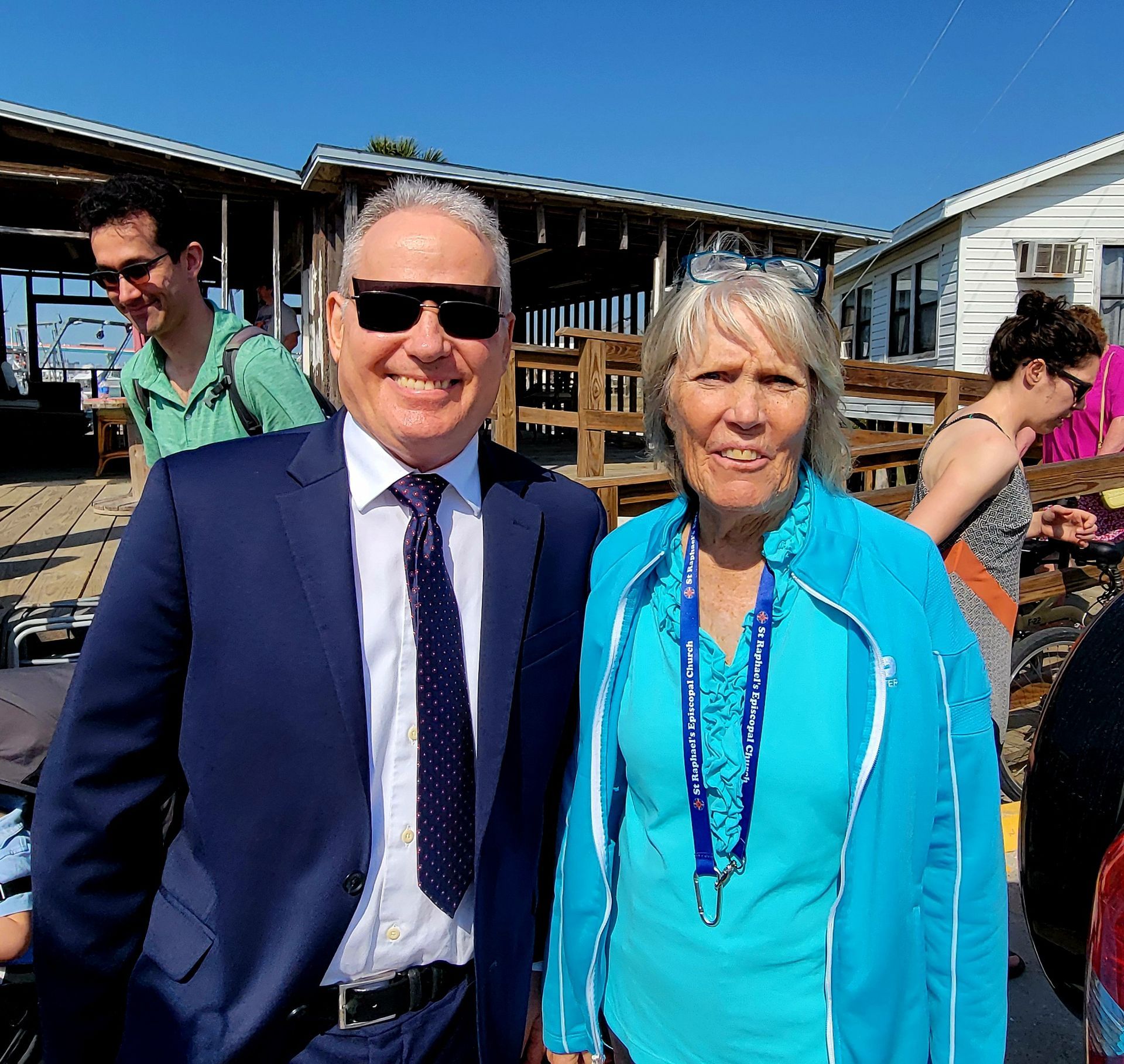 Man in suit and woman in blue pose outdoors, smiling, with others in background.
