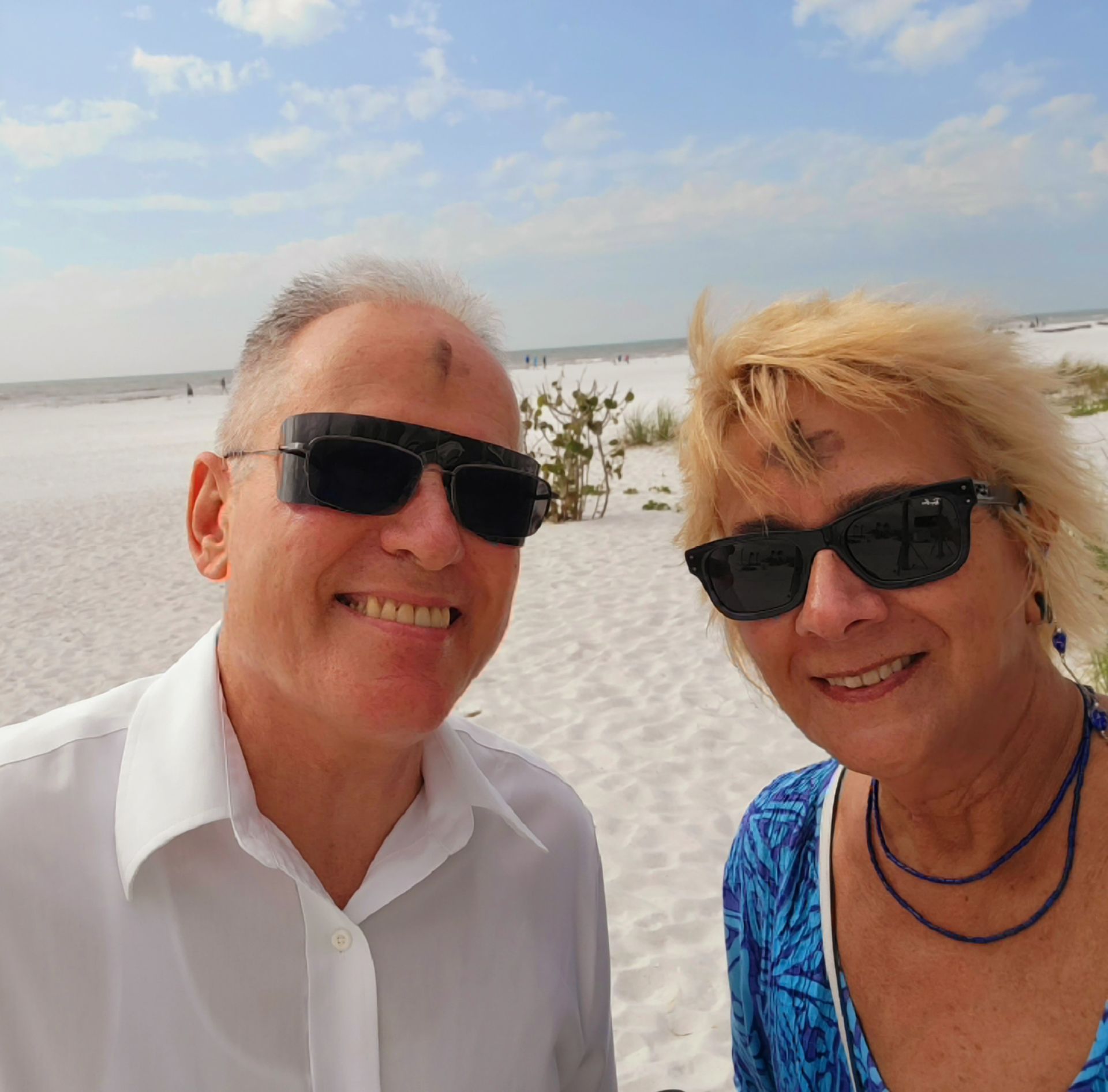 Smiling couple on a white sand beach with ashes on their foreheads, wearing sunglasses.