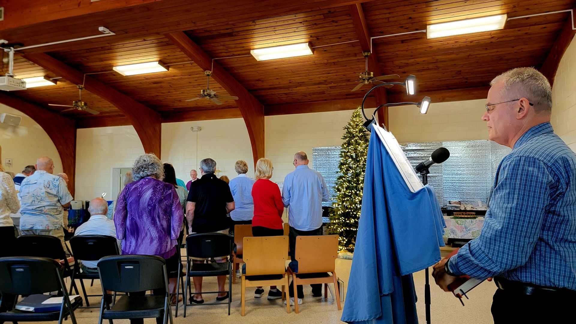 People stand in a room, facing a Christmas tree; a man speaks from a podium.
