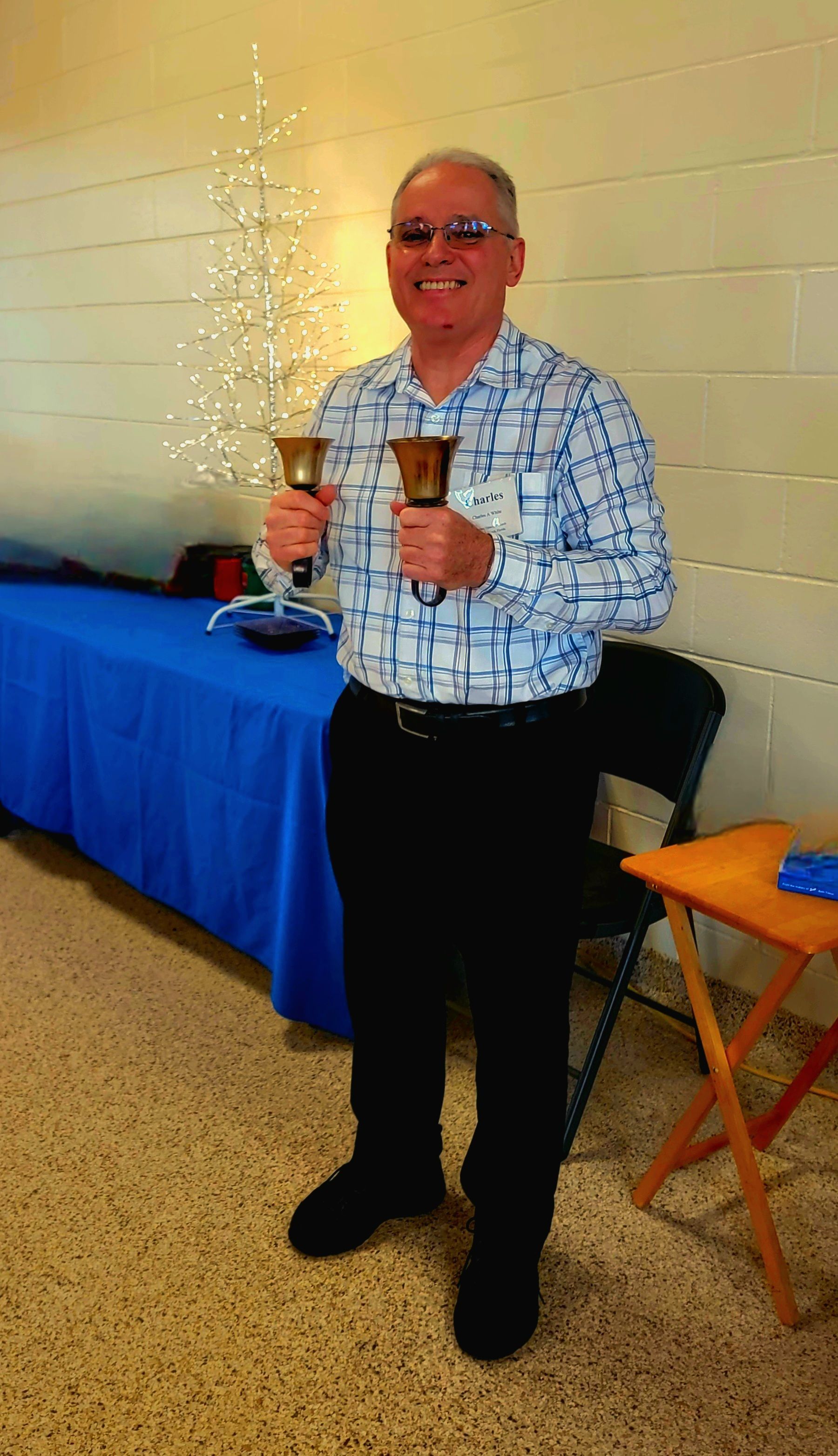 Man smiling, holding two handbells. He wears glasses, a blue patterned shirt, black pants, in a room.