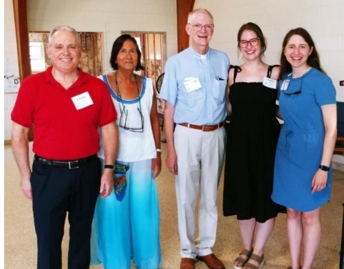 Five people smiling for a photo. A man in a red shirt stands to the left, and a woman in blue pants is on the right.