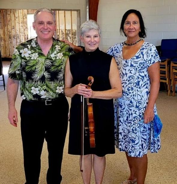 Three people pose for a photo indoors; a man in a floral shirt, a woman with a violin, and another woman in a blue dress.