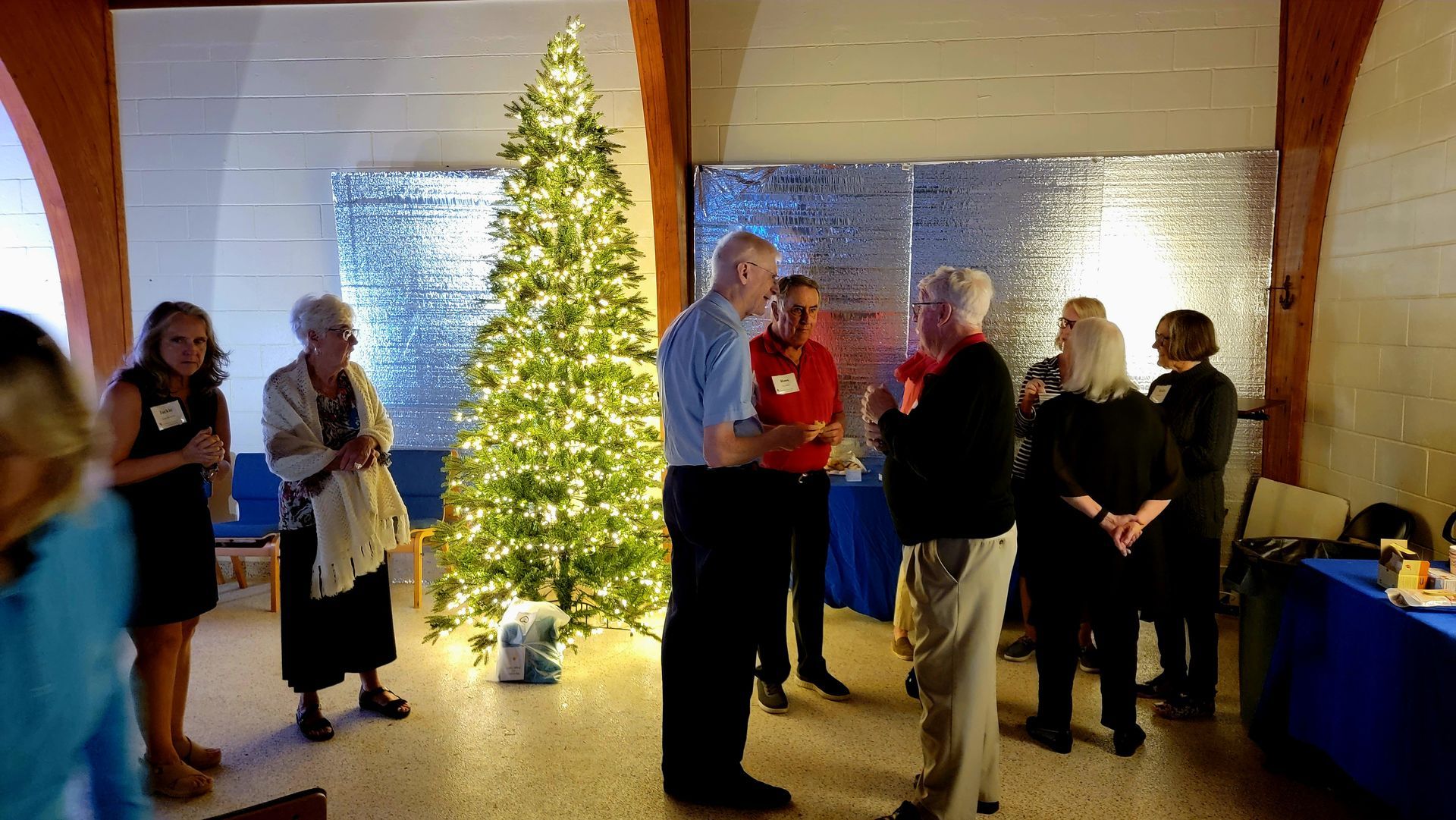 People gather near a lit Christmas tree, socializing in a room with a blue table and silver wall.