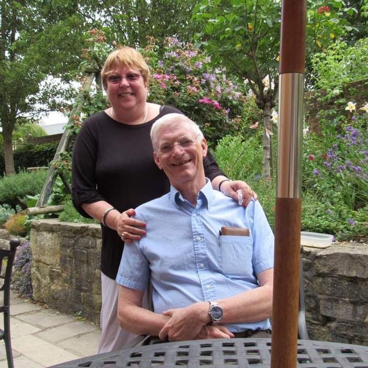 Woman with her hands on the shoulders of a smiling older man, seated. Outdoors near greenery.