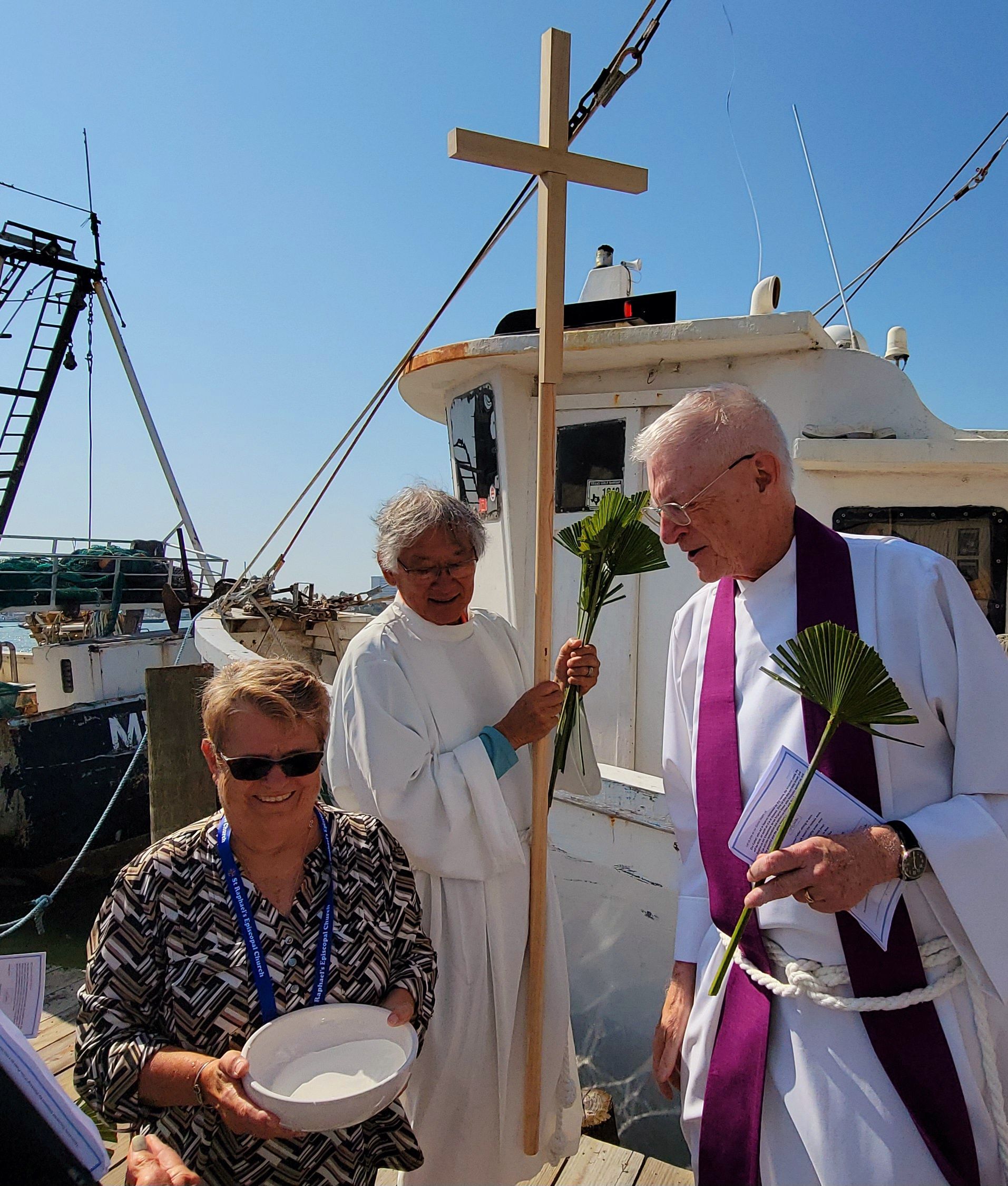 Clergy bless a boat at a dock. People with cross and holy water, blue sky.