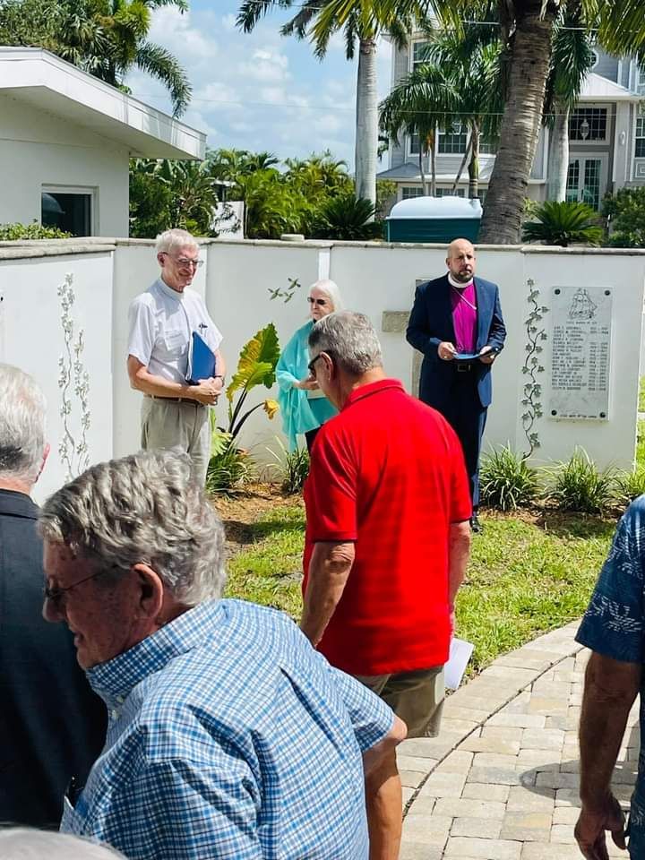 Group of people gathered outside; a man in a red shirt is in the foreground, others listen to a speaker in a suit.