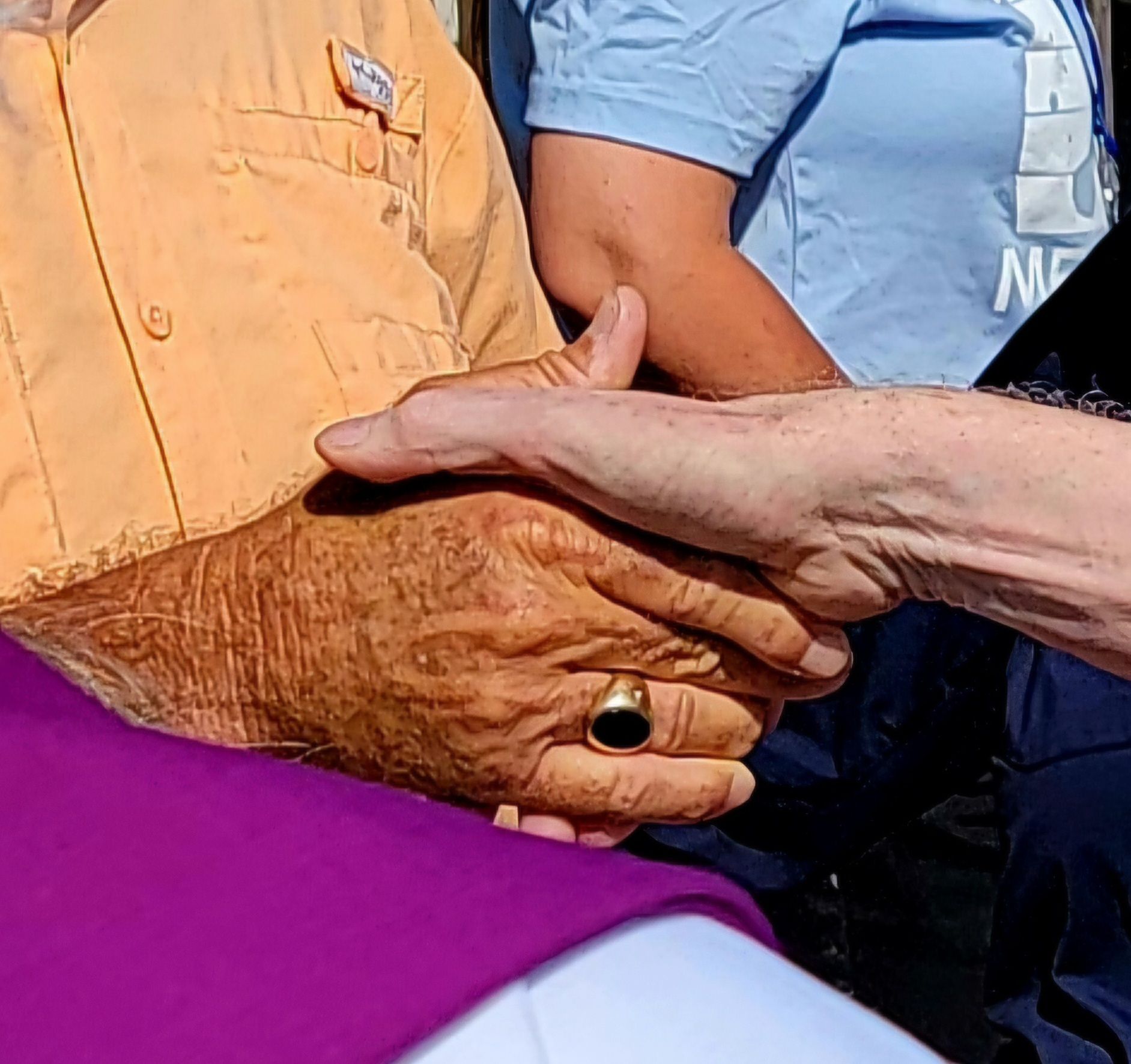 Two people clasp hands. One wears a gold ring, the other a purple robe. People wearing blue shirts are behind.