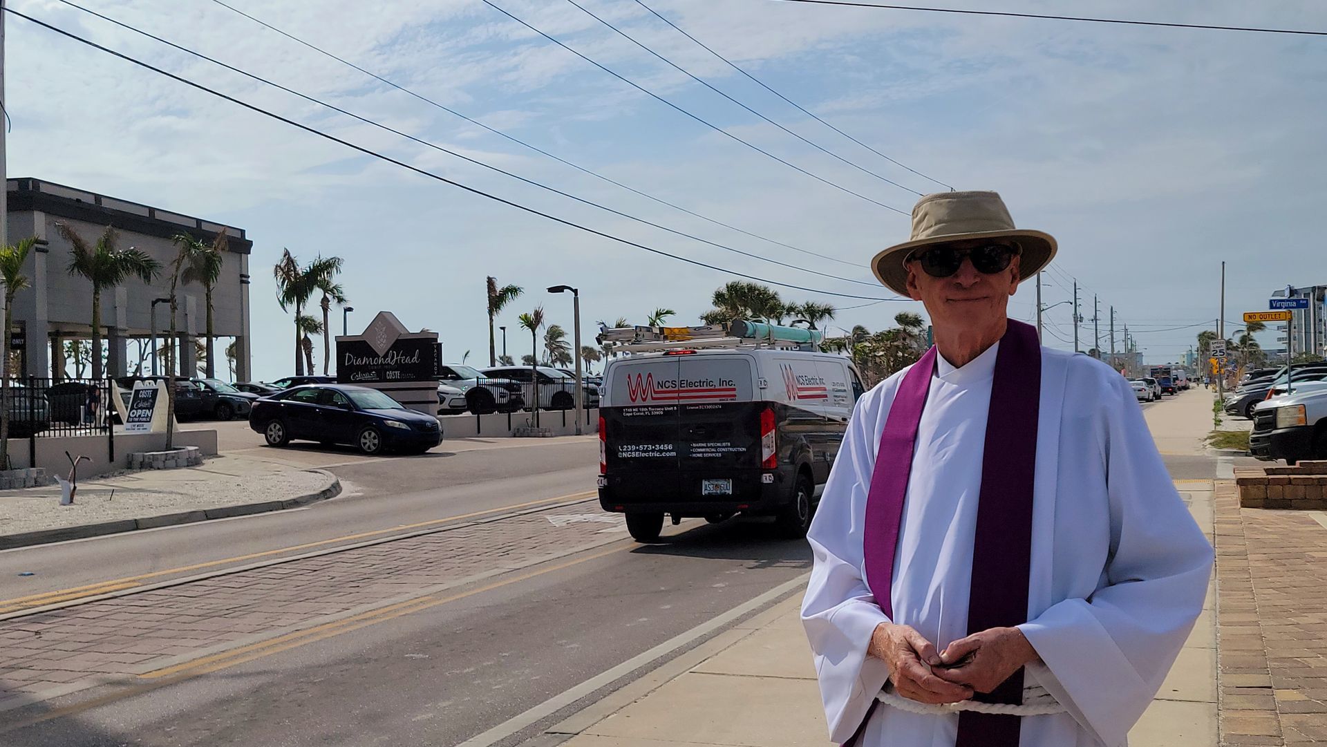 Man in clerical robes stands on a street, holding hands, with cars and ocean in background.