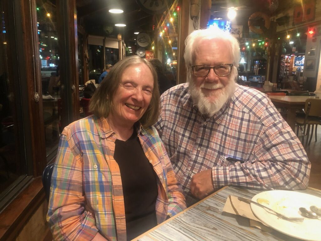 Couple at a restaurant, smiling. Man has a white beard, wearing glasses, checkered shirt.