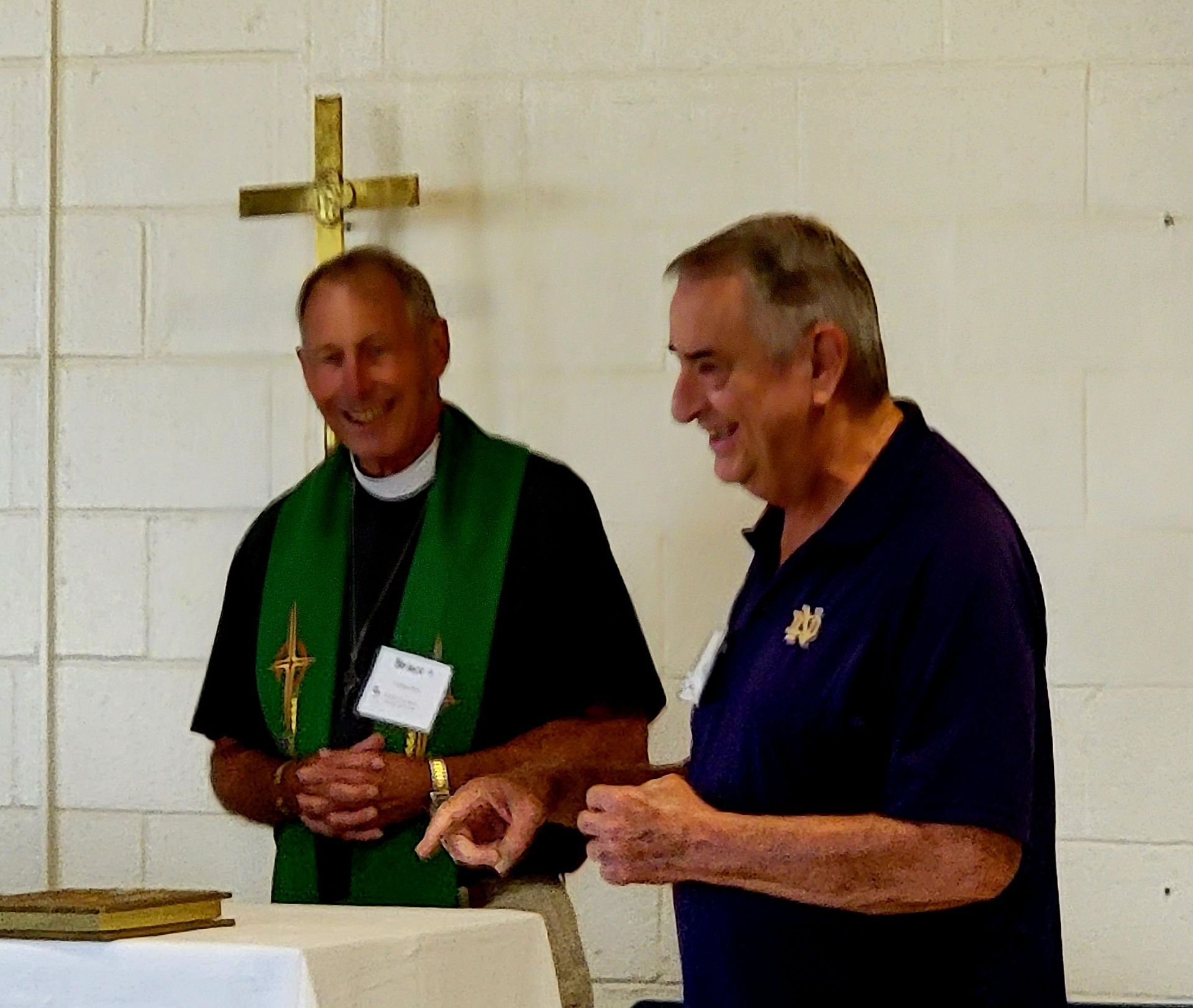 Two men laugh, one a priest with green stole, the other in a blue shirt