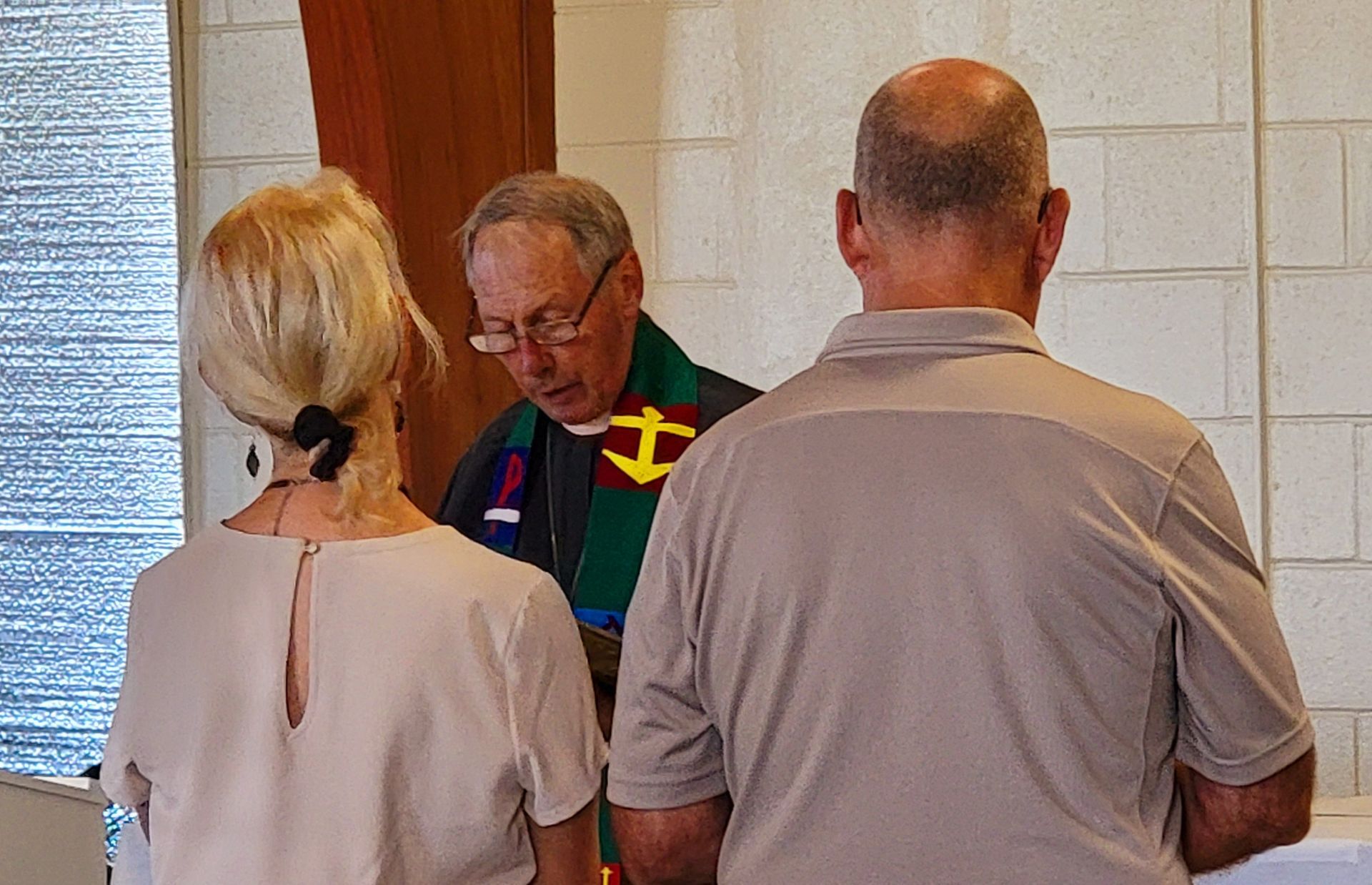 Couple at altar, priest officiating. Blonde woman, man with bald head, officiant in robe. Ceremony setting.