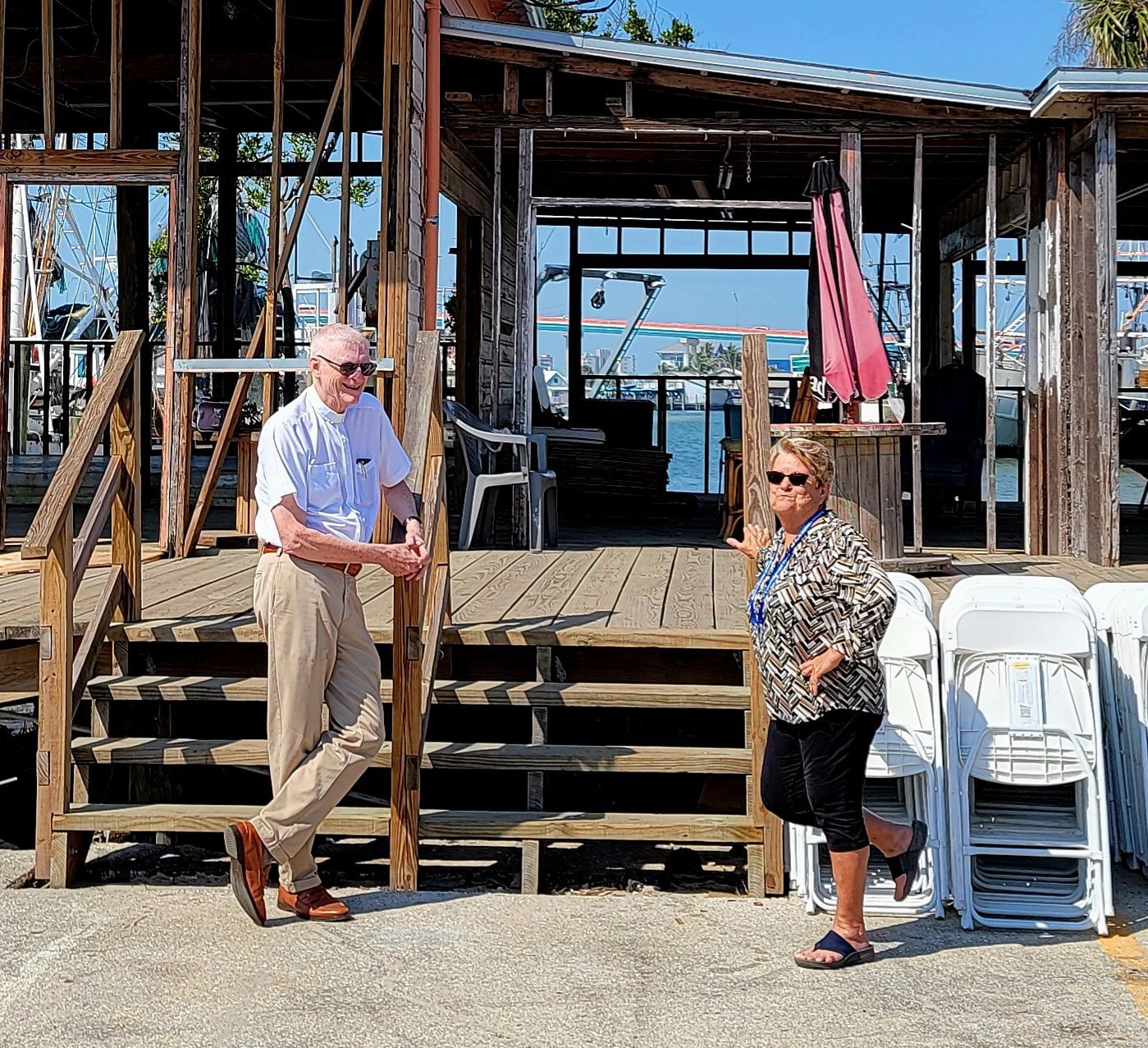 Man and woman standing on a weathered wooden deck, overlooking water. They pose near a partially built structure.